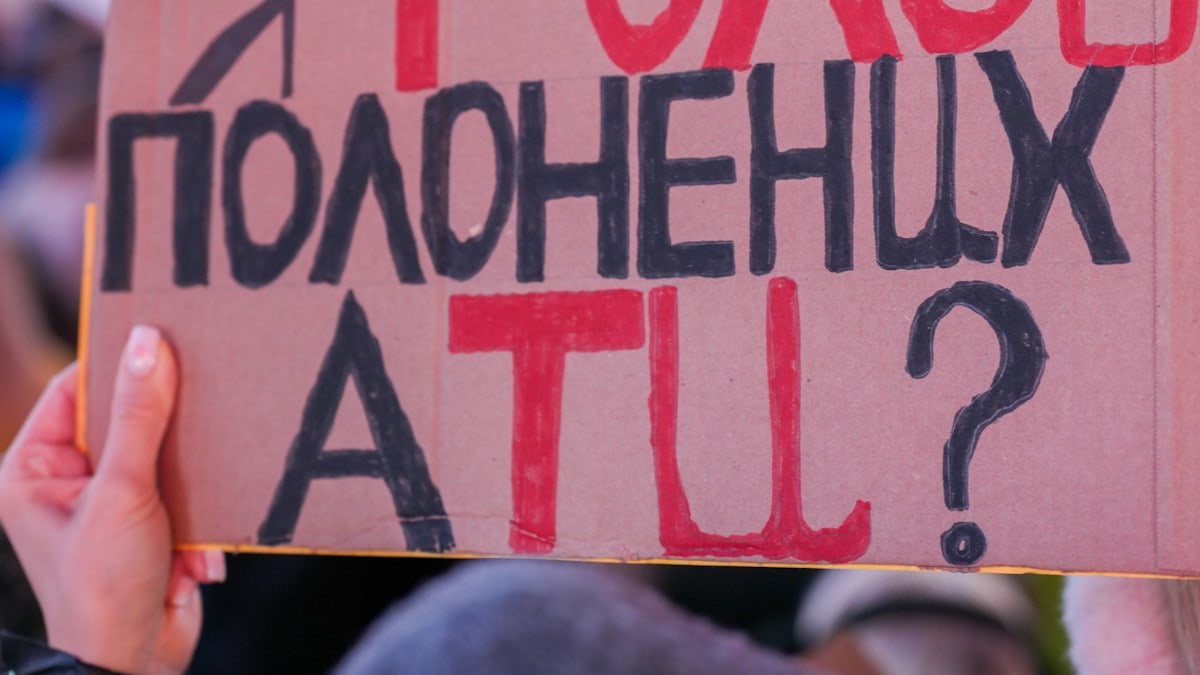 Close-up of a hand holding a handmade cardboard protest sign lettered in red and black Ukrainian text asking who will be the voice of captives. The crowd behind is softly blurred, with hints of blue and yellow flags at a nighttime anti-war rally. Protesters rally in Times Square, New York City, on February 15, 2022, standing in solidarity with Ukraine and speaking out against the war at an anti-war demonstration. Photo: Olek Buzunov (@buzunov.photoart)
